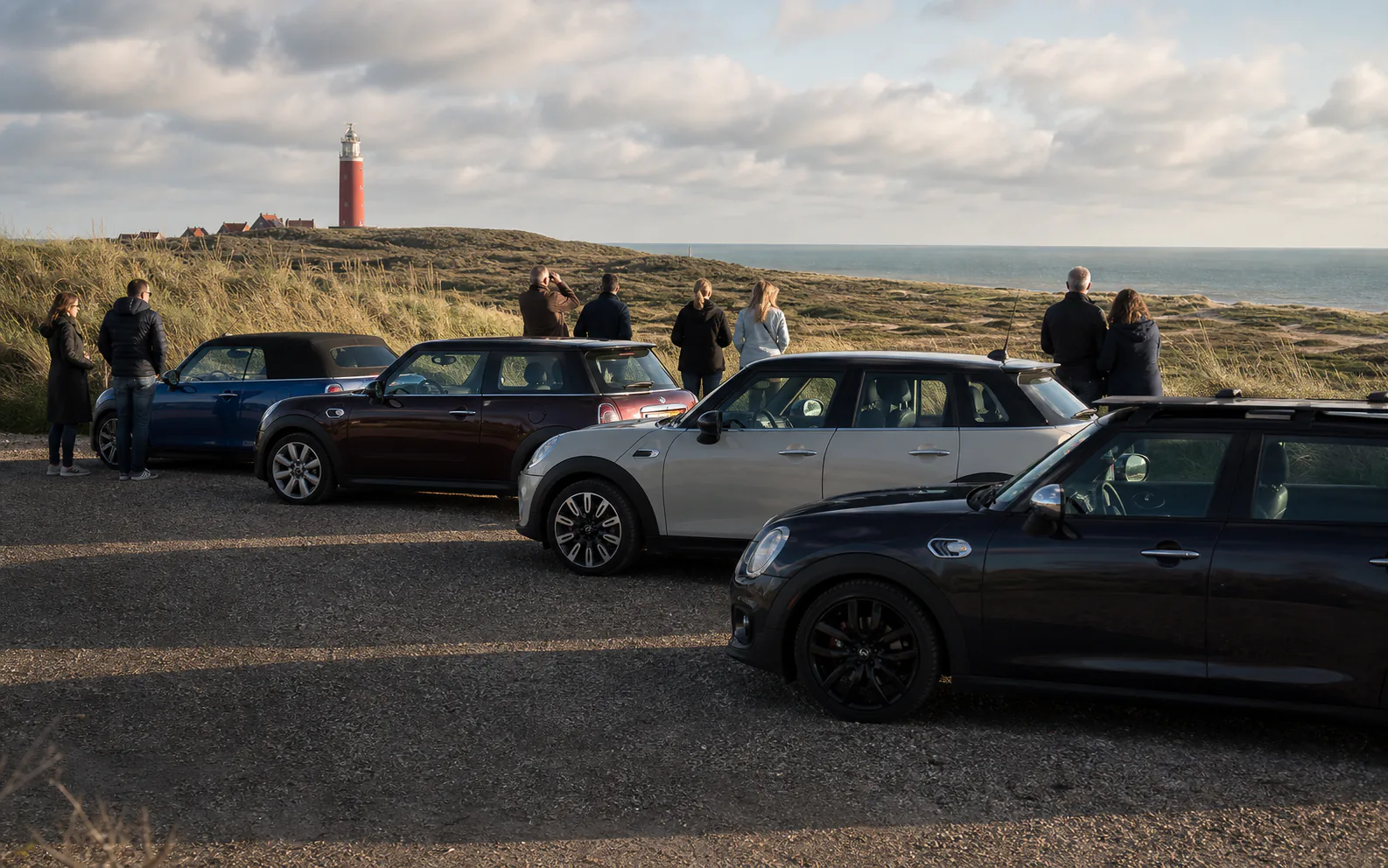 Deelnemers maken foto's bij geparkeerde MINI's met de rode vuurtoren en Texelse duinen in de verte